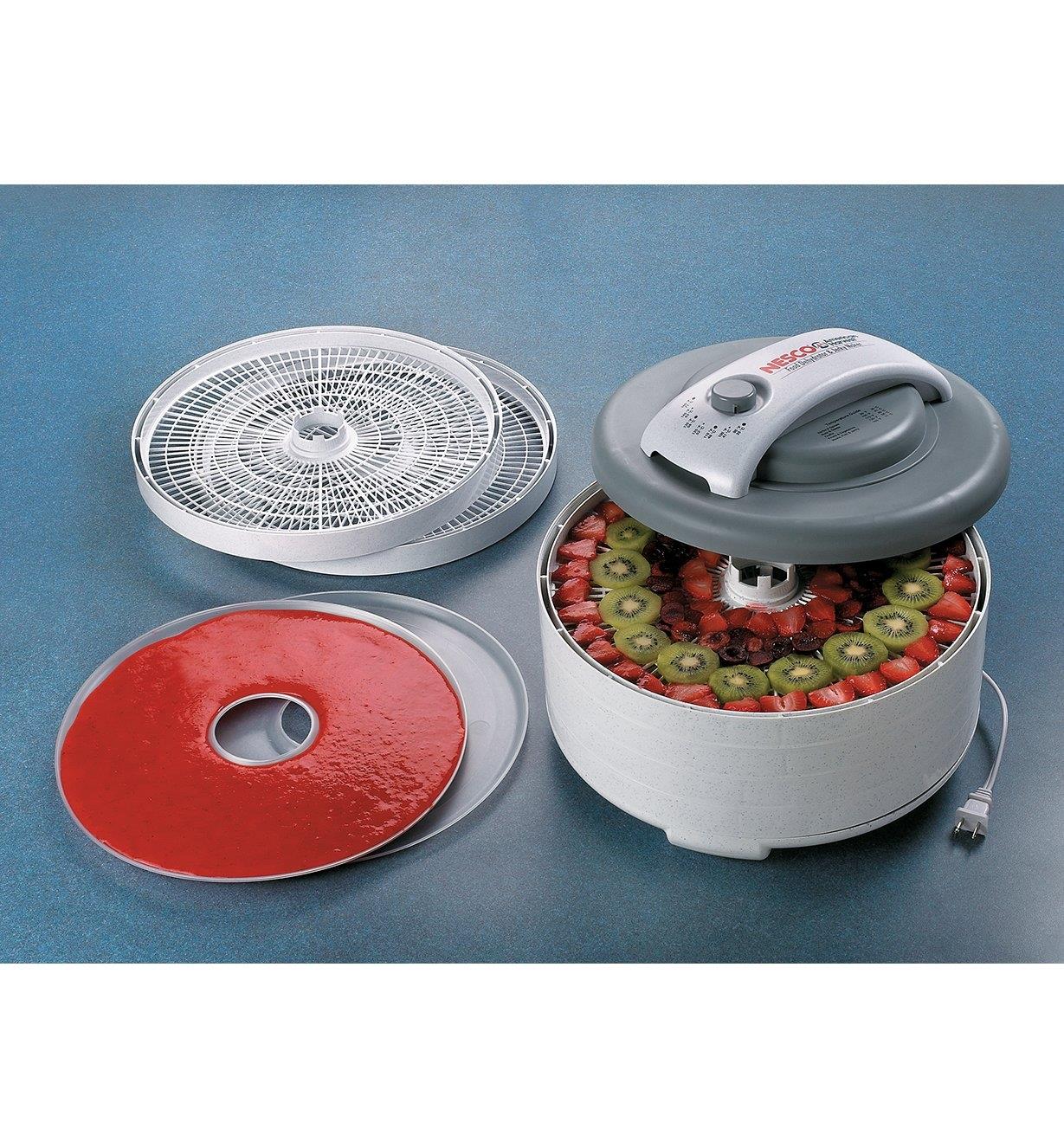 Food Dehydrator containing fresh fruit slices, with a filled fruit roll sheet and extra trays sitting beside it