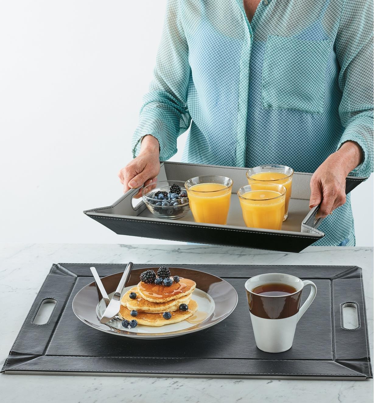 Femme transportant des verres de jus d'orange et un bol de petits fruits dans un plateau à rebord pliant FreeForm et plateau déposé à plat sur un comptoir avec une assiette de crêpes et une tasse de thé à l'avant-plan