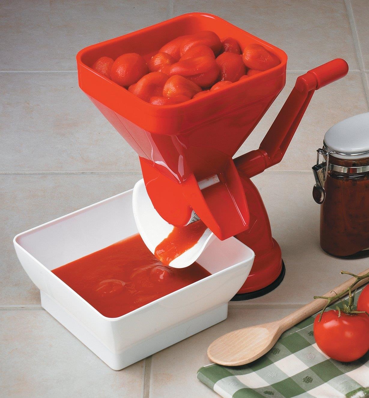 Tomato press sits on a counter with the hopper full of whole tomatoes and pulp running into the catch bowl