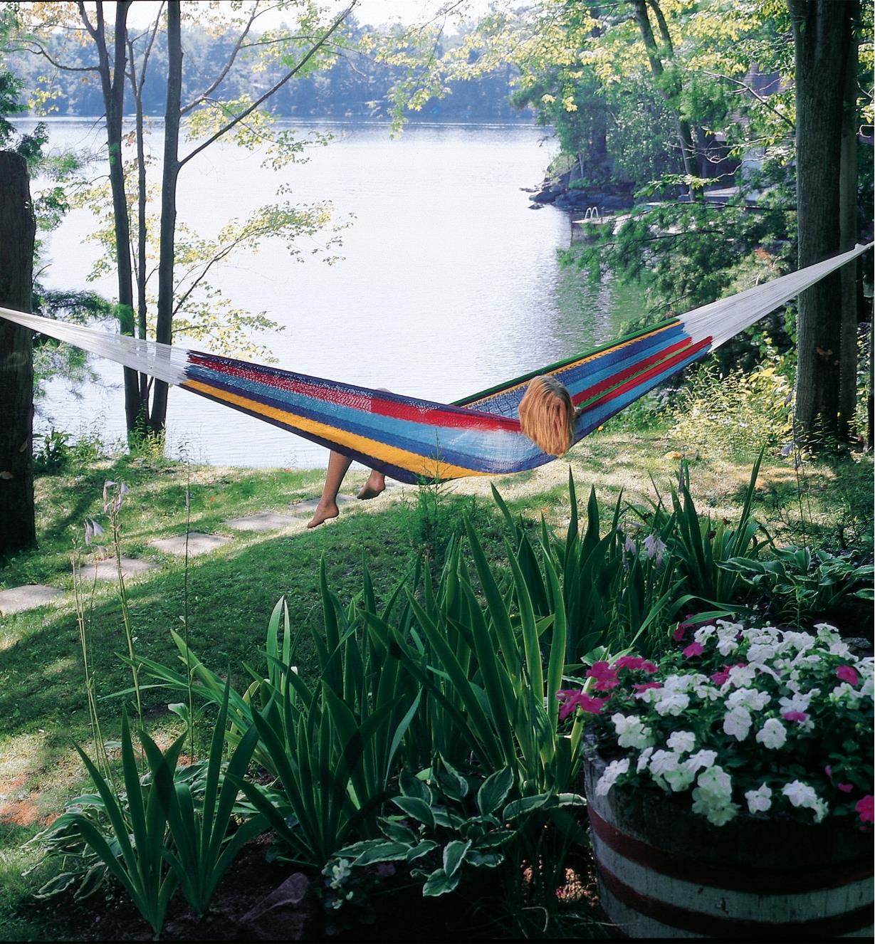 A person lies crossways on a Mayan Hammock hung between trees by a lake