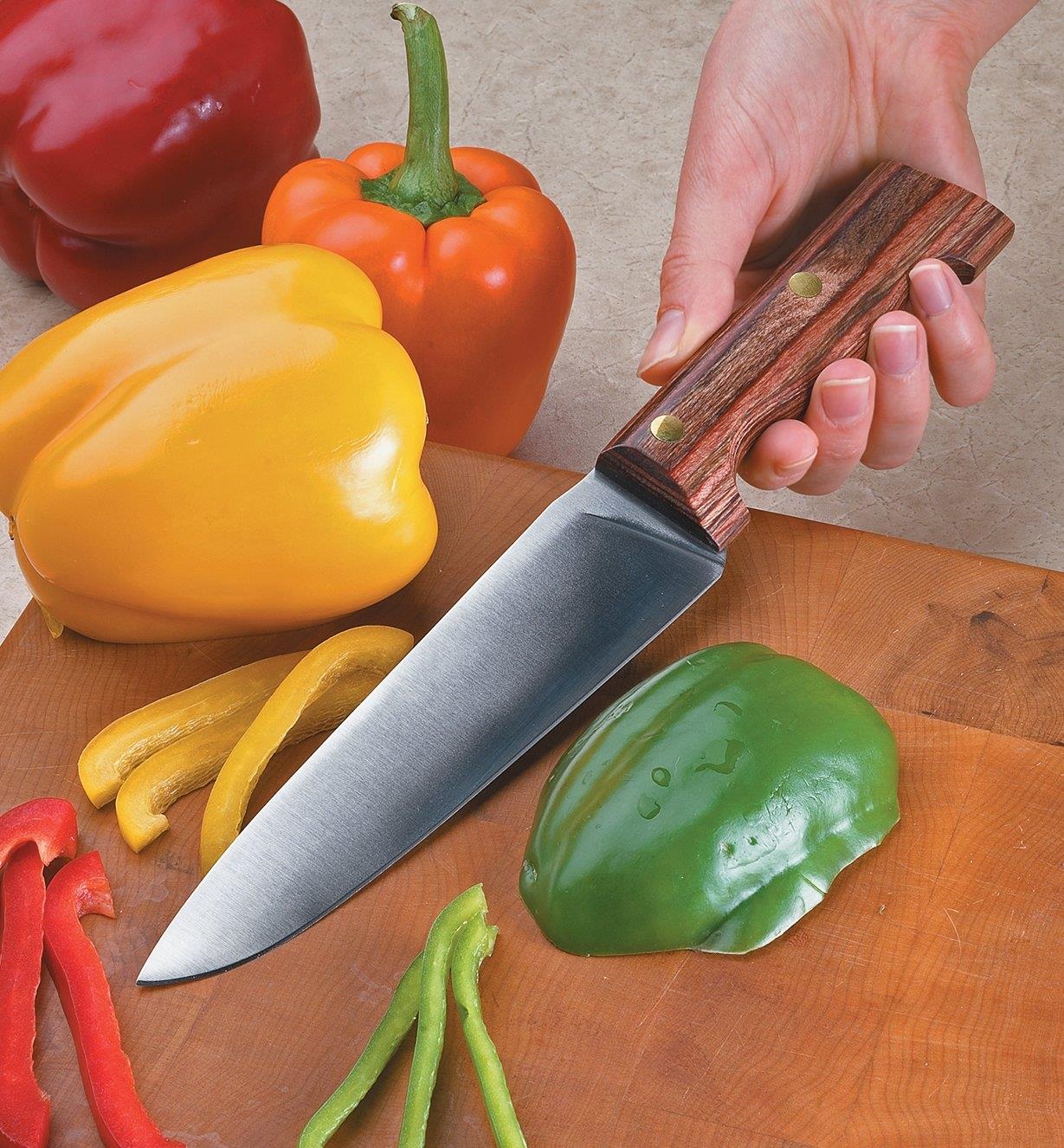 Personne tenant un couteau de chef campagnard appuyé contre une planche à découper près de poivrons de couleurs variées