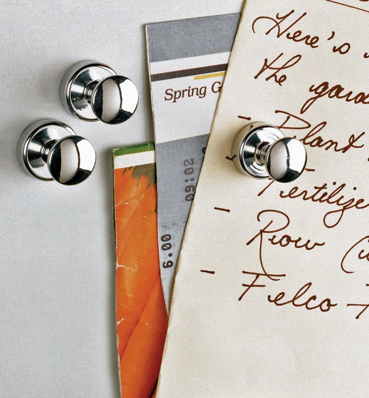 Three fridge magnets on a fridge, one holding up a grocery list and other paper items
