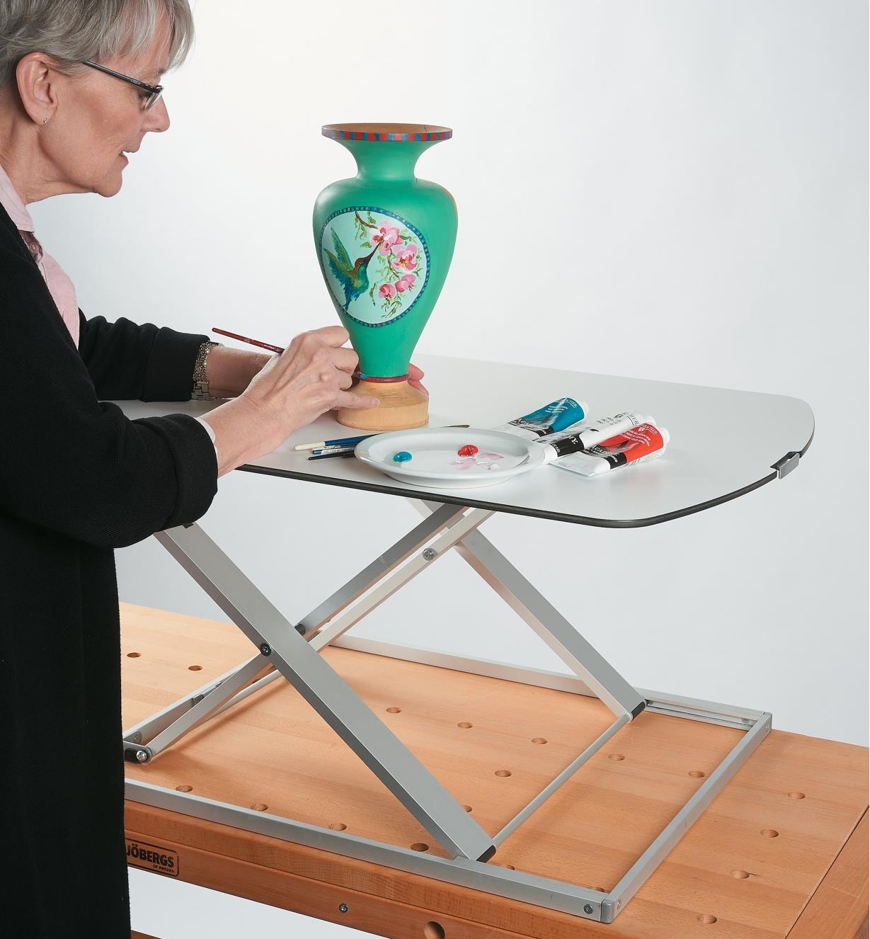 A woman paints a vase on the Height-Adjustable Work Stand