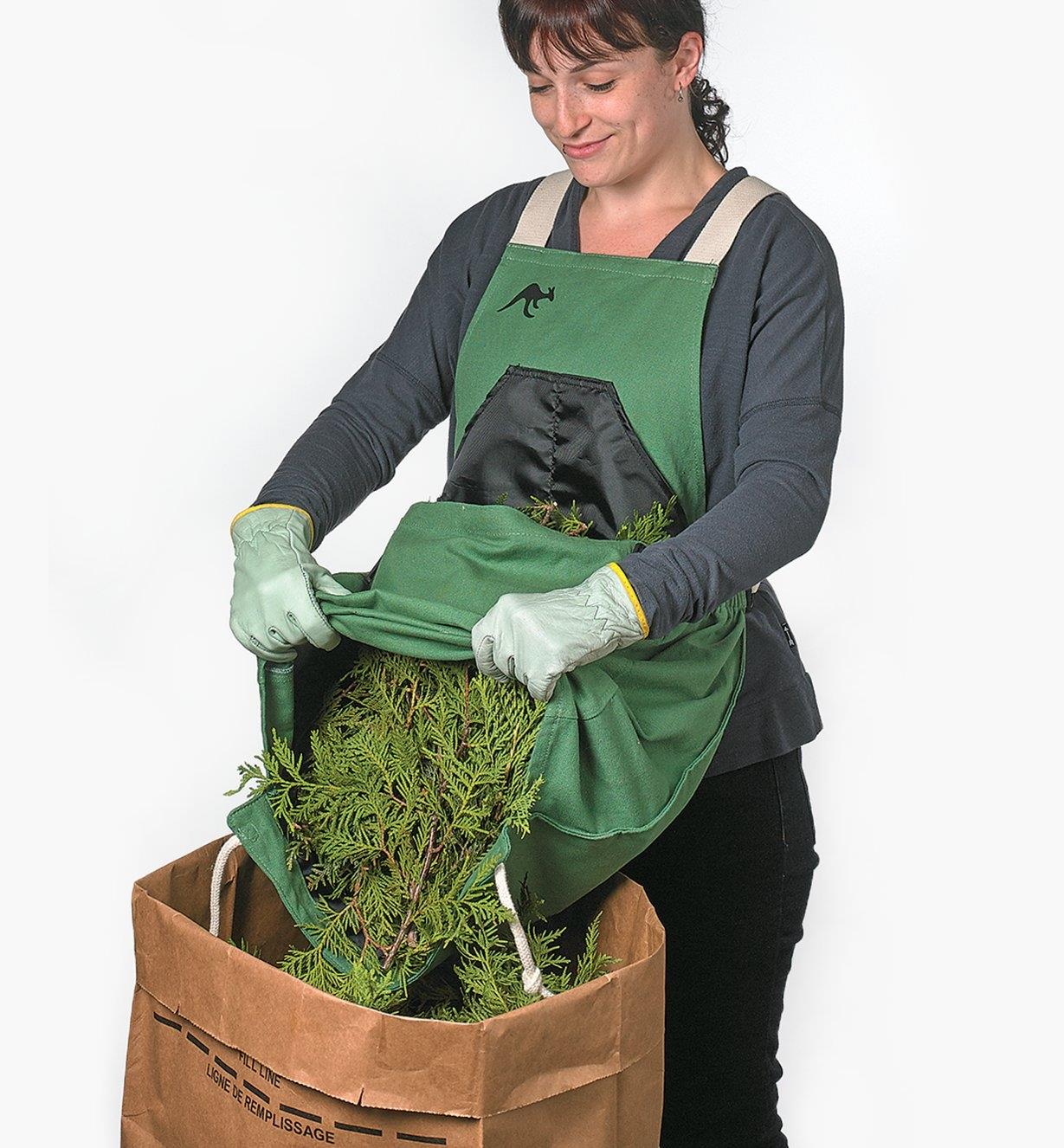 A woman wearing the kangaroo pocket apron dumps cedar trimmings from the pocket into a paper bag