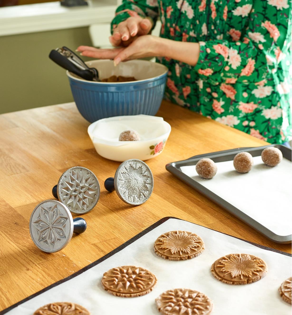 Three cookie stamps sitting next to a tray of cookies that have been stamped, while a woman in the background rolls more cookie dough