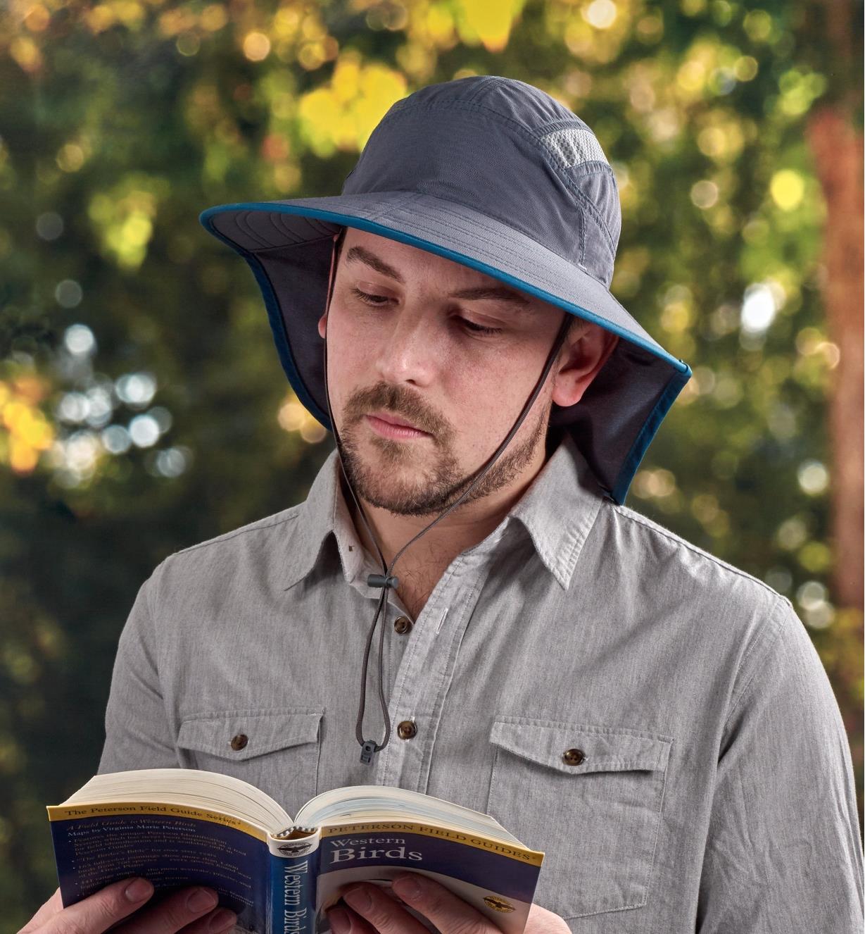 A man wearing an adventure sun hat reads a book outdoors