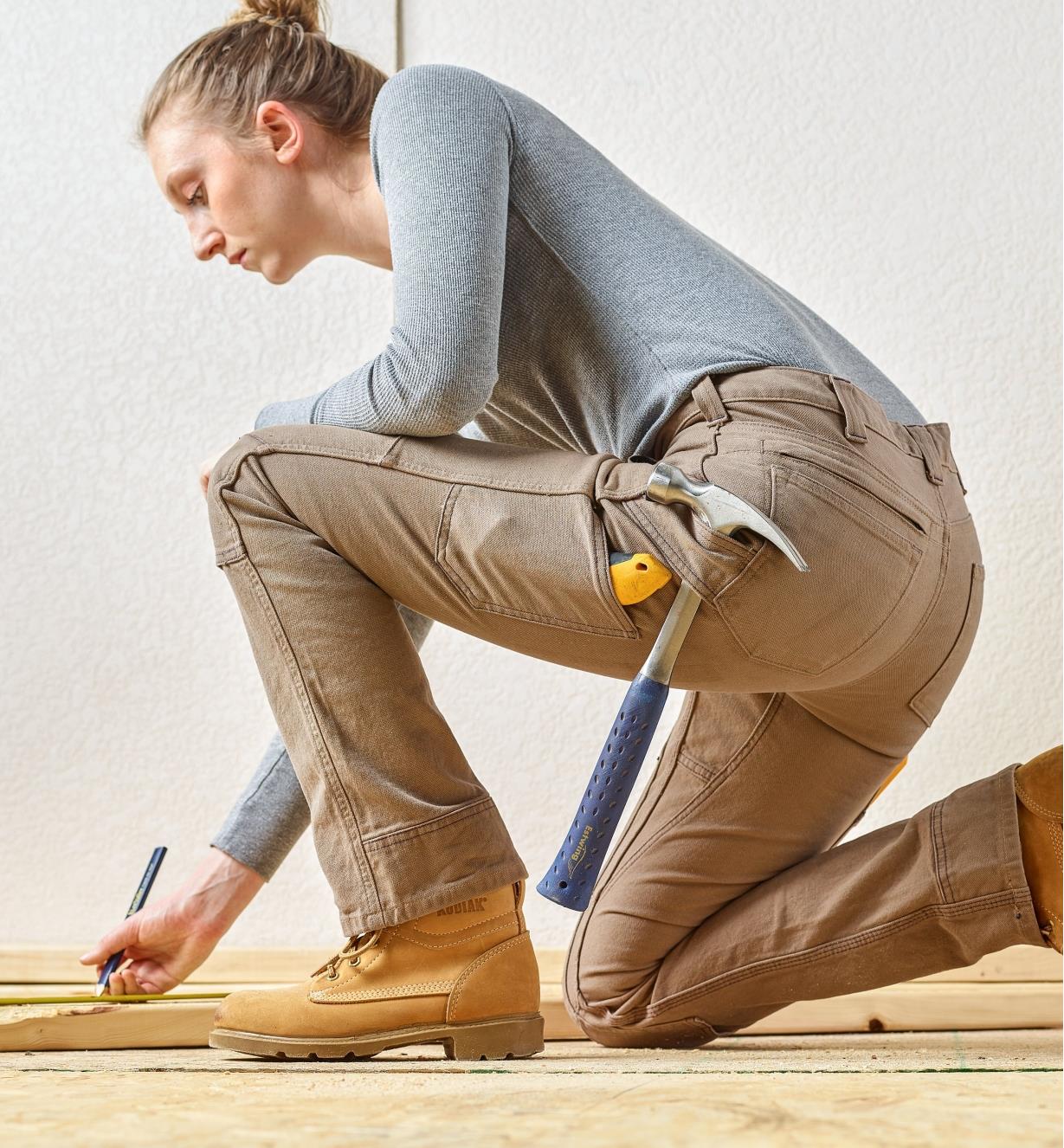 A woman wearing Dovetail pants bends on one knee to  mark a board