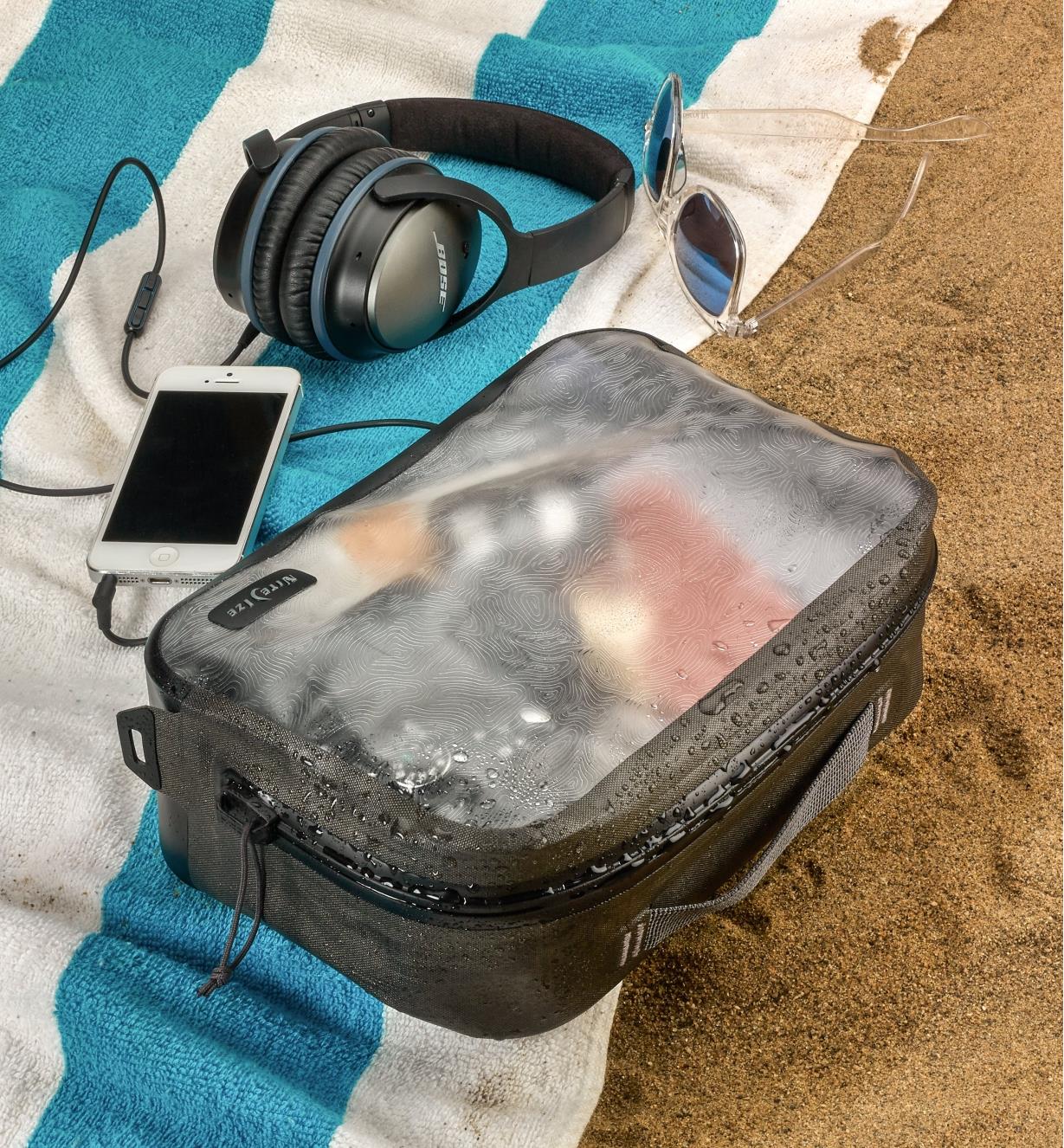 A closed RunOff medium packing cube filled with items lies on a beach towel with a cell phone, headphones and sunglasses nearby