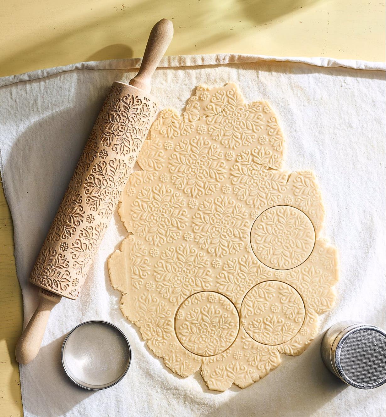 An embossing rolling pin on a pastry cloth beside embossed raw dough cut with circular cookie cutters