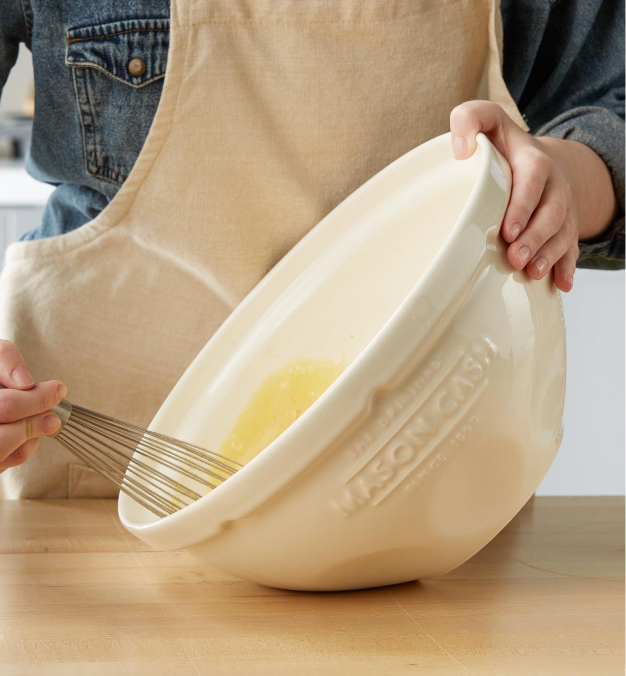 A cook who is whisking eggs tilts a Mason Cash mixing bowl to rest on a flat area of its side