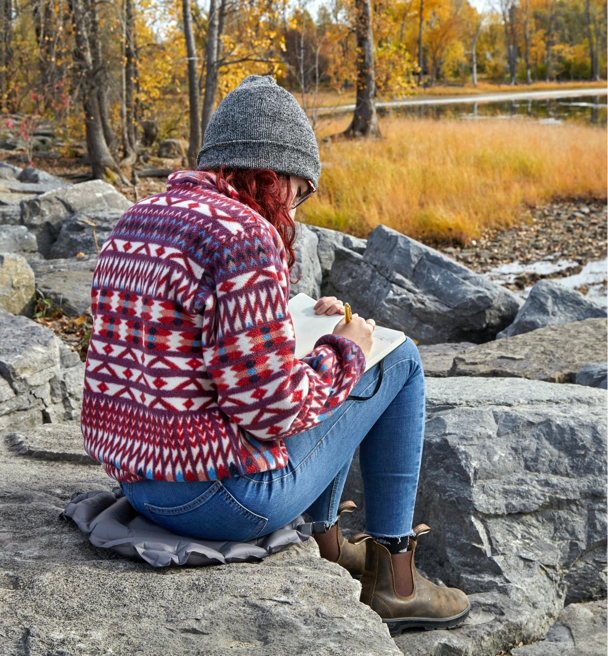 A woman sits on an inflatable seat cushion placed on a rock while writing notes outdoors