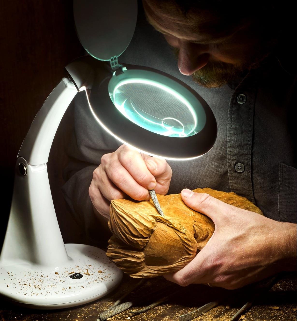 A man uses an LED tabletop magnifying lamp to help carve a piece of wood