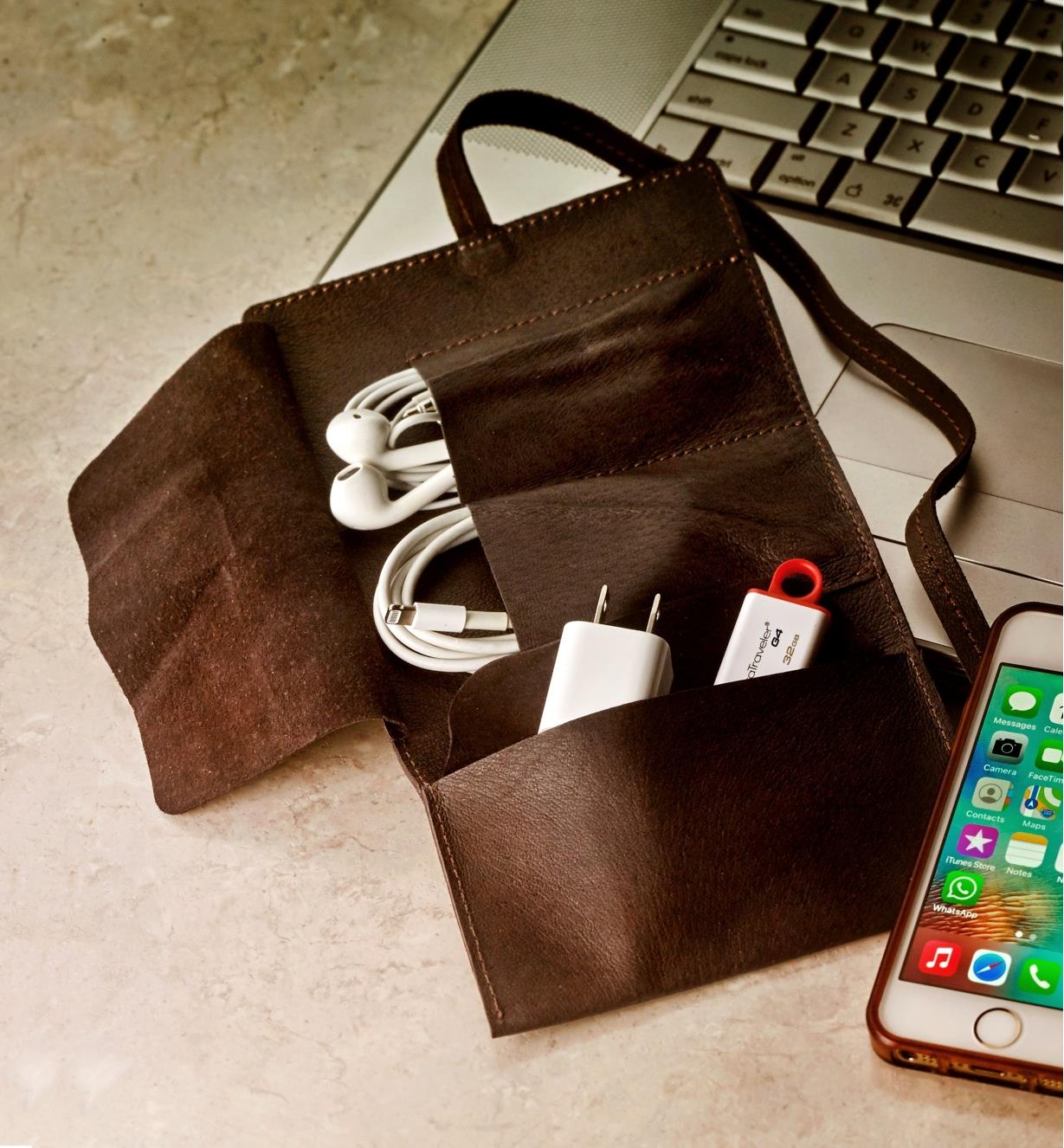 A cord wallet beside a laptop and smartphone, holding a charge cable, ear buds and a flash drive