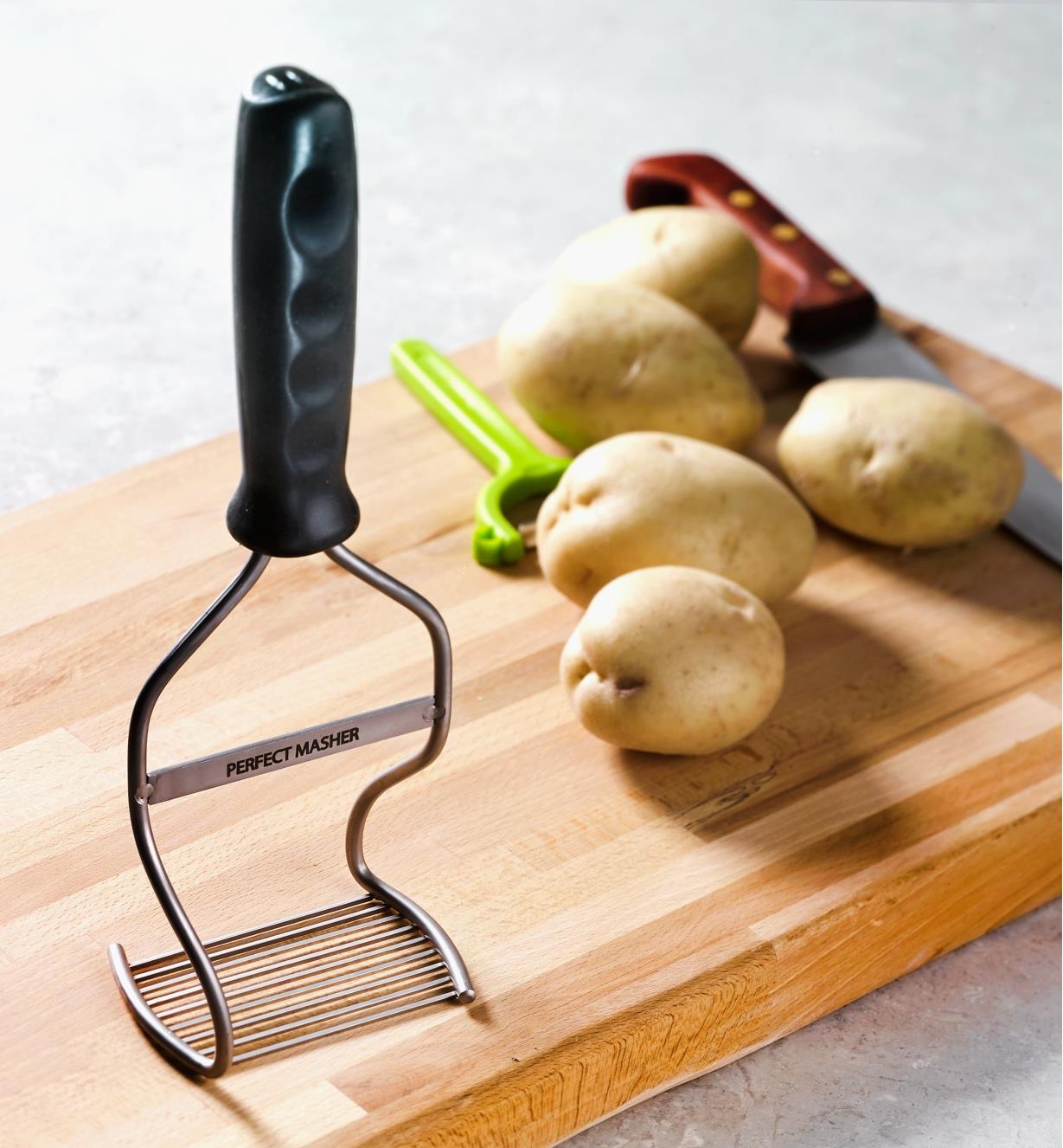 Potato Masher placed on a cutting board next to potatoes, a peeler and a knife