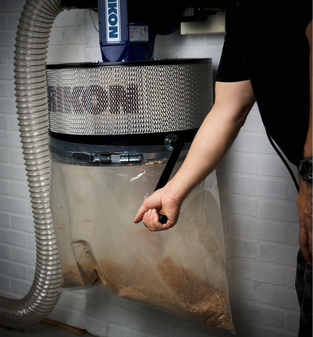 A person cranking the handle of a Rikon Wall-Mount Dust Collector with the dust bag attached to the bottom
