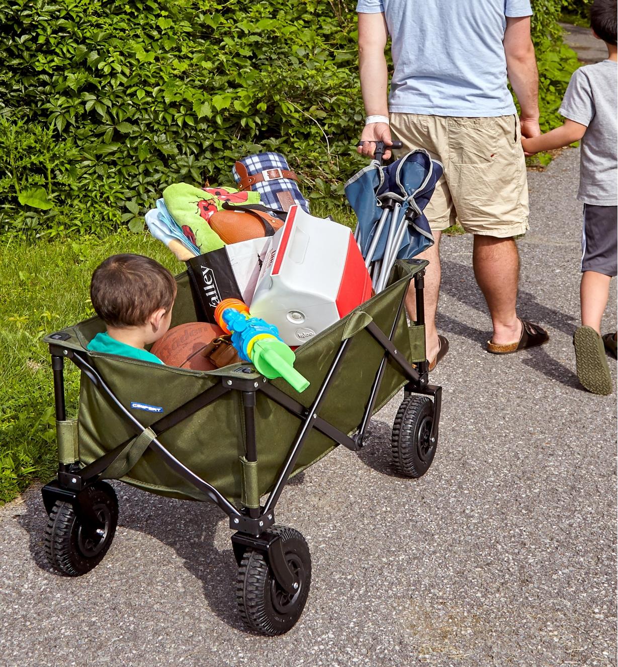 A father pulls a boy in a folding cart, along with a cooler and other supplies for a day in the park