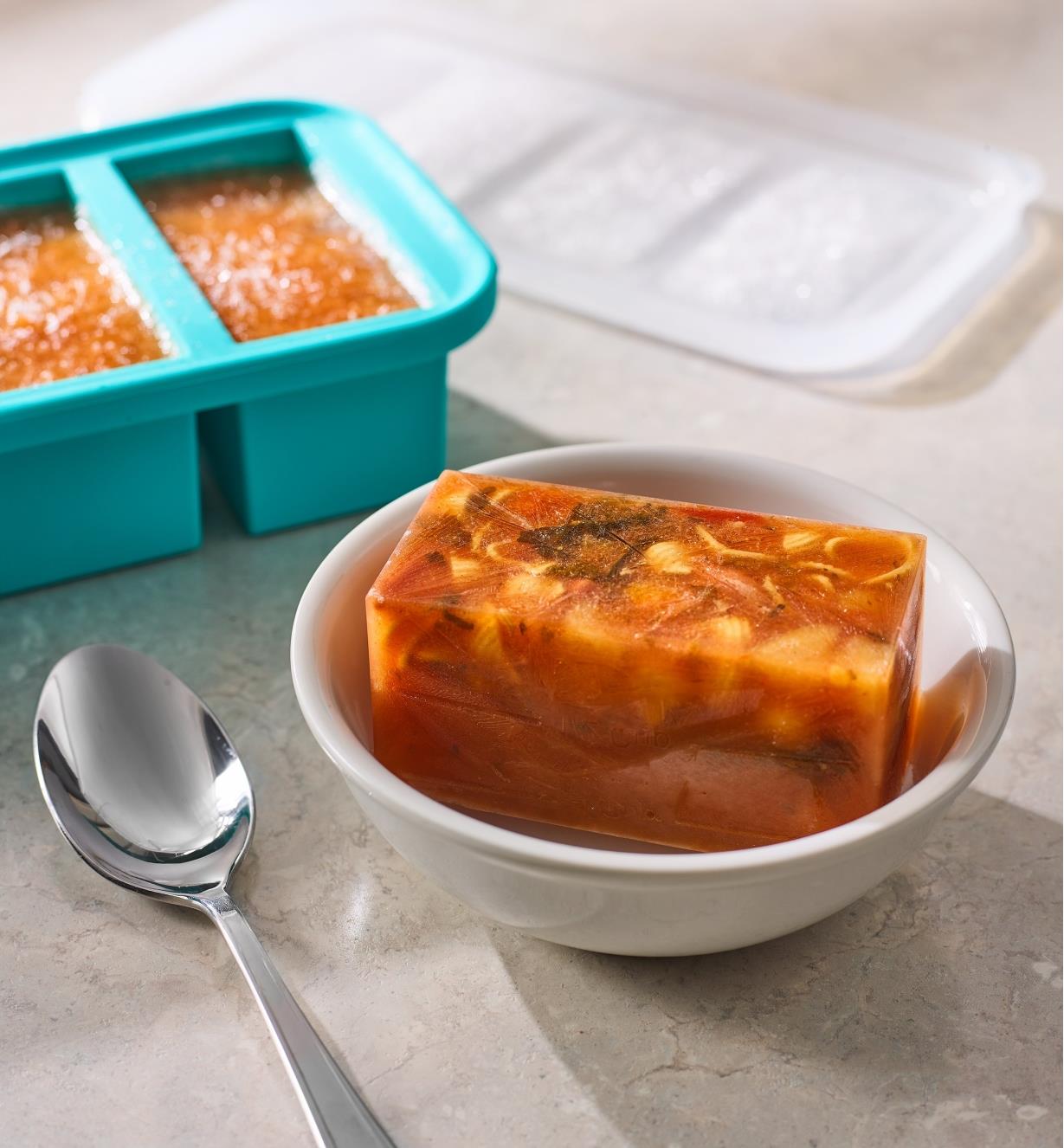 A block of frozen soup in a bowl with a Souper Cube tray behind it