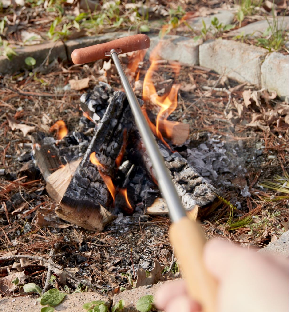 Personne faisant griller une saucisse sur un feu de camp à l'aide d'une fourchette télescopique pour feu de camp