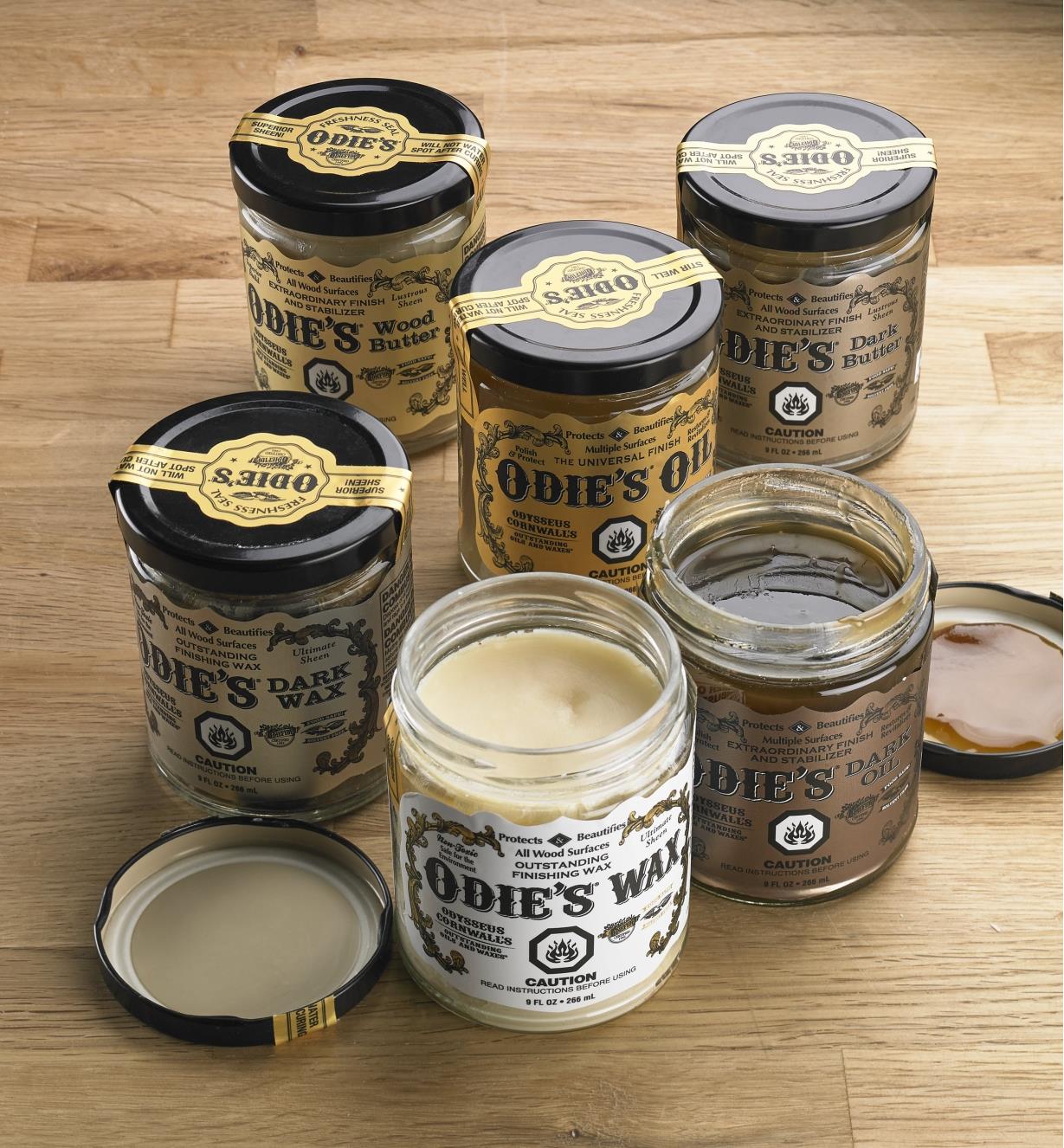All six Odie’s Oil Finishes are on a wood surface, with two of the jars open