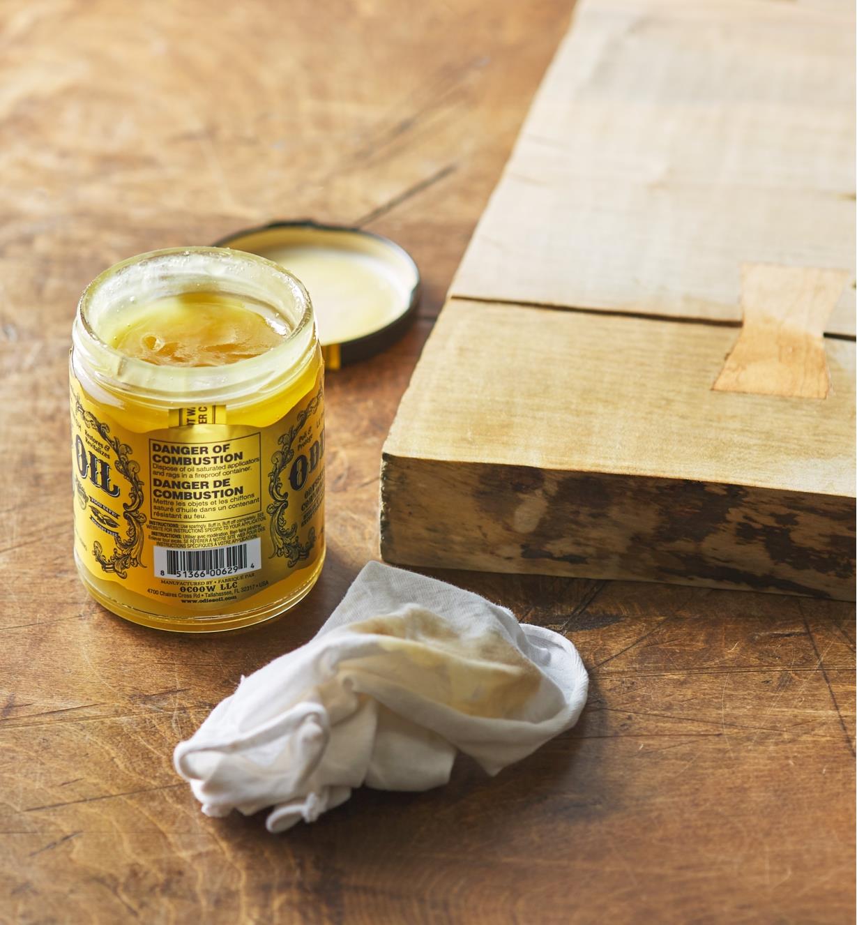 An open jar of Odie’s Oil next to a white cloth and a half-treated plank of wood