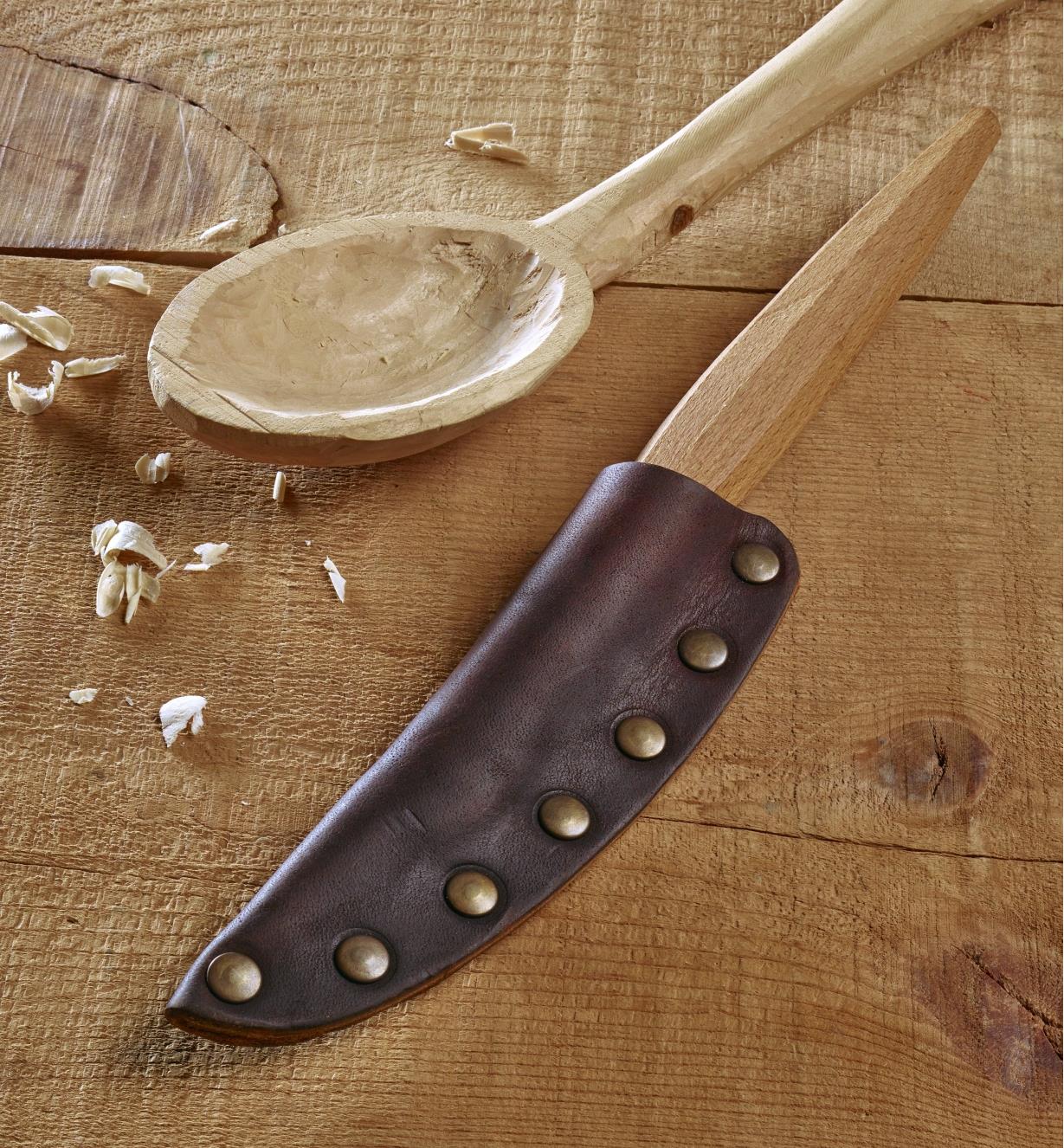 The leather knife sheath attached to a spoon carving knife next to a wooden spoon
