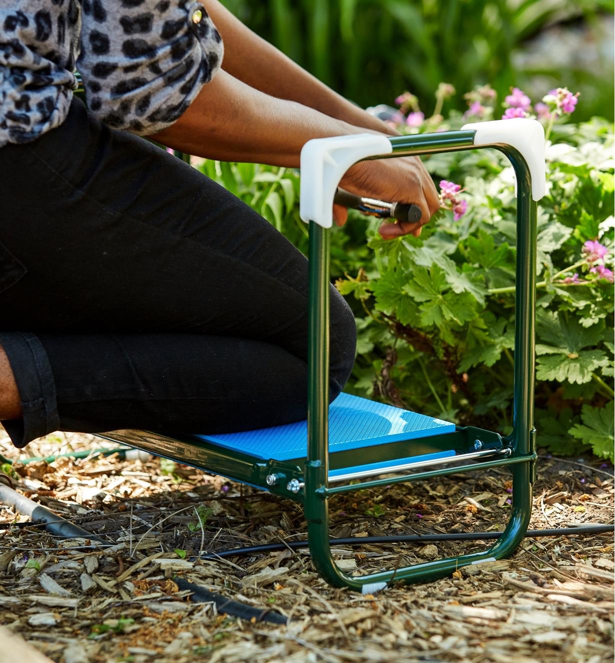 A woman kneels on the folding kneeler stool to tend to garden flowers growing low to the ground