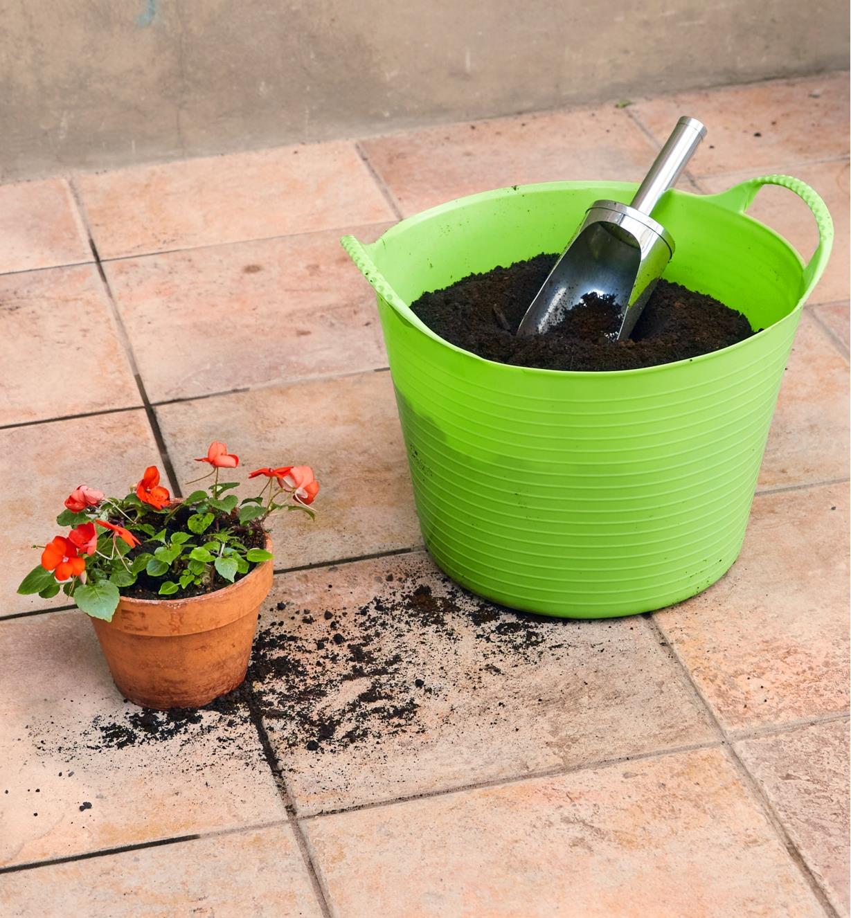 Des impatientes fraîchement plantées dans un pot posé sur un plancher carrelé près d’un panier de jardin de 14 L rempli de terreau