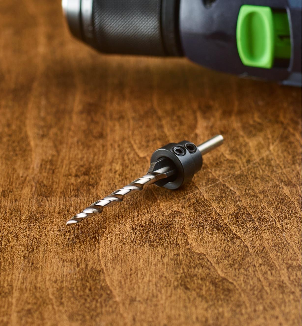 A drill bit with a stop collar sits in front of a drill on a table