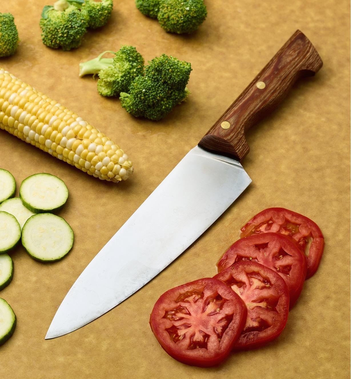 Grand couteau de chef à côté de rondelles de tomates et de courgettes, d’un épi de maïs et de fleurons de brocoli