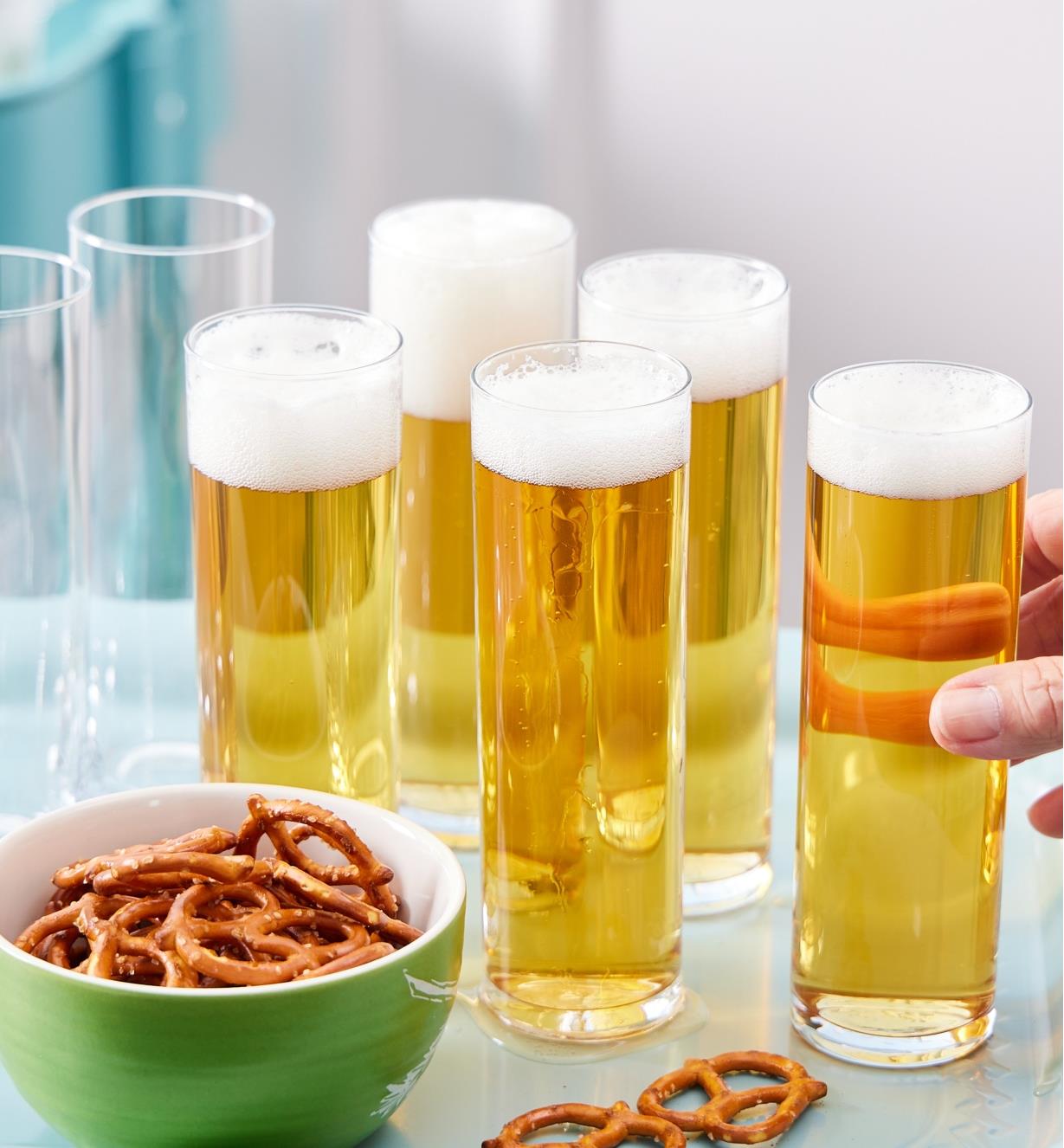 Five Kölsch glasses filled with beer next to a bowl of pretzels