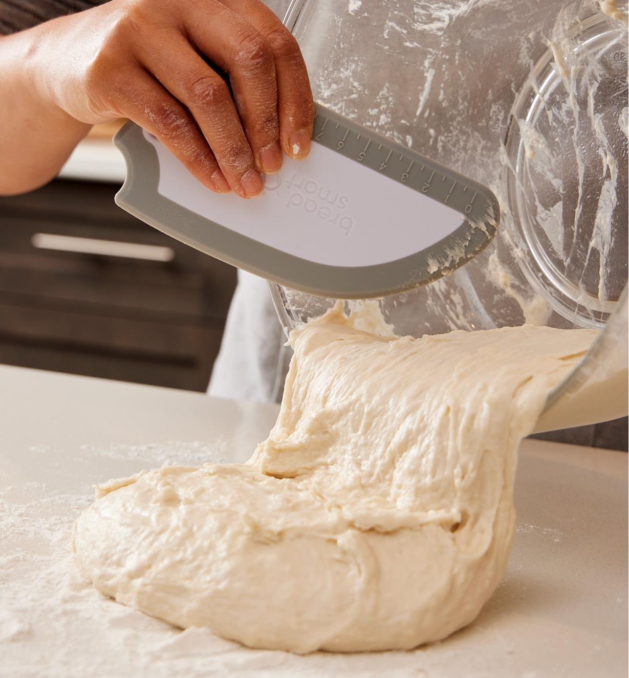 Placing fresh dough on a floured countertop, using a bowl scraper to remove it from a mixing bowl