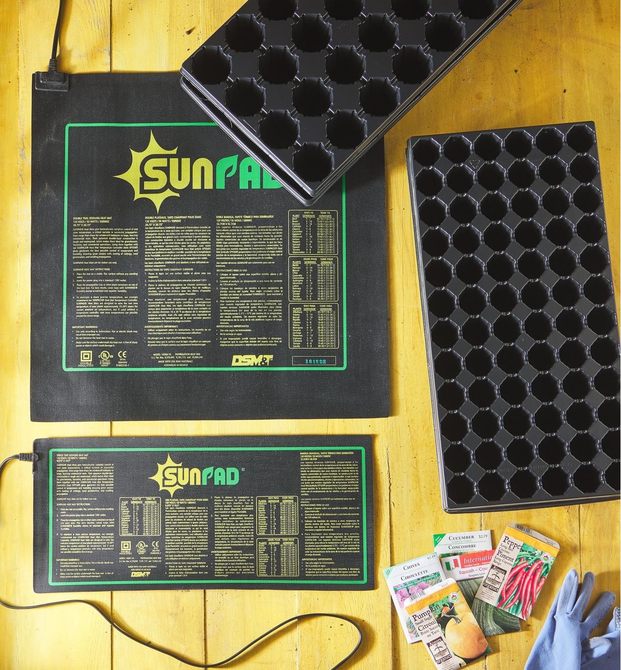 Two sizes of seedling heat mats shown next to empty seed trays, seed packets and gardening gloves