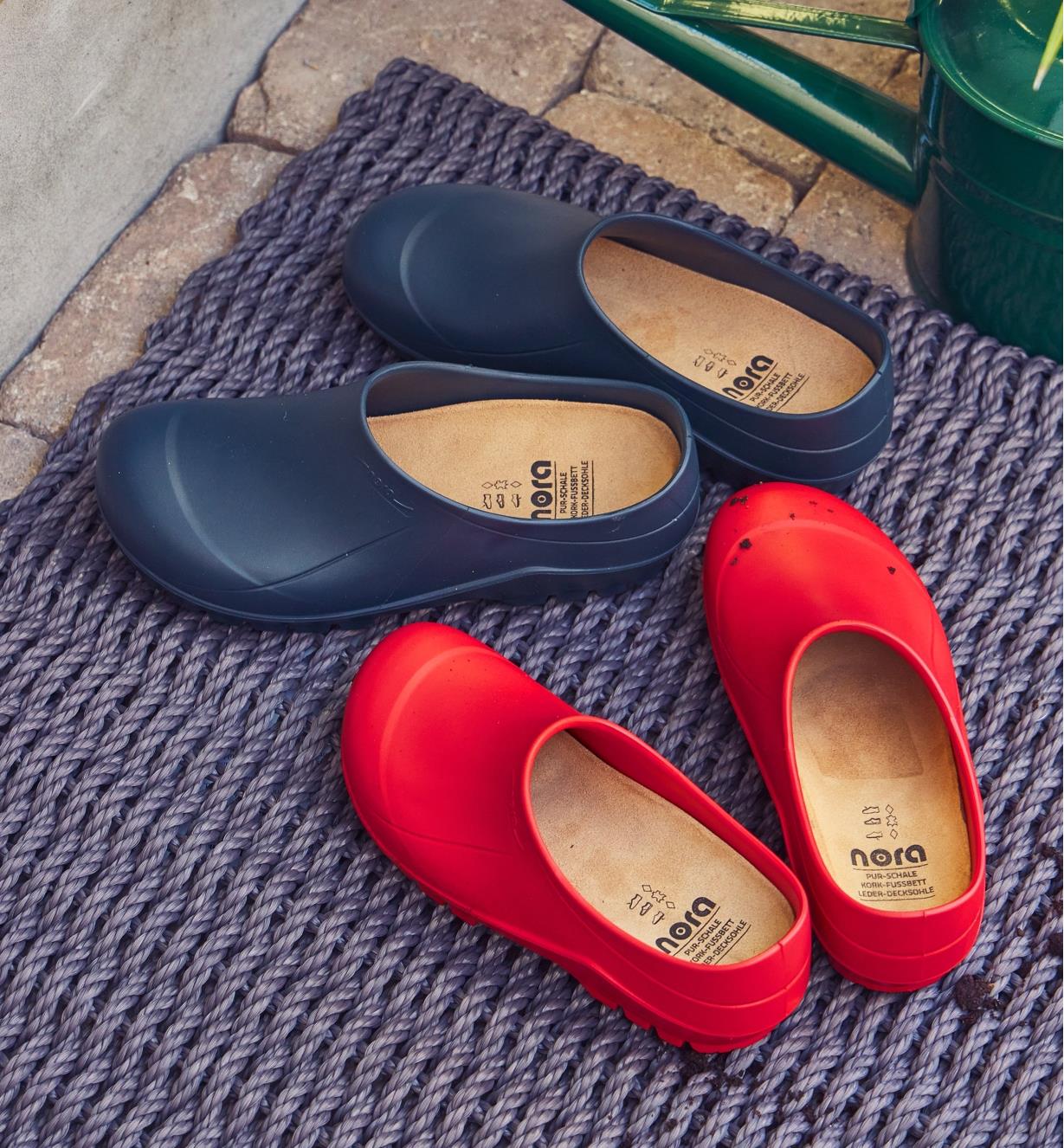 Two pairs of European garden clogs, one red and one navy, placed on a doormat on a patio