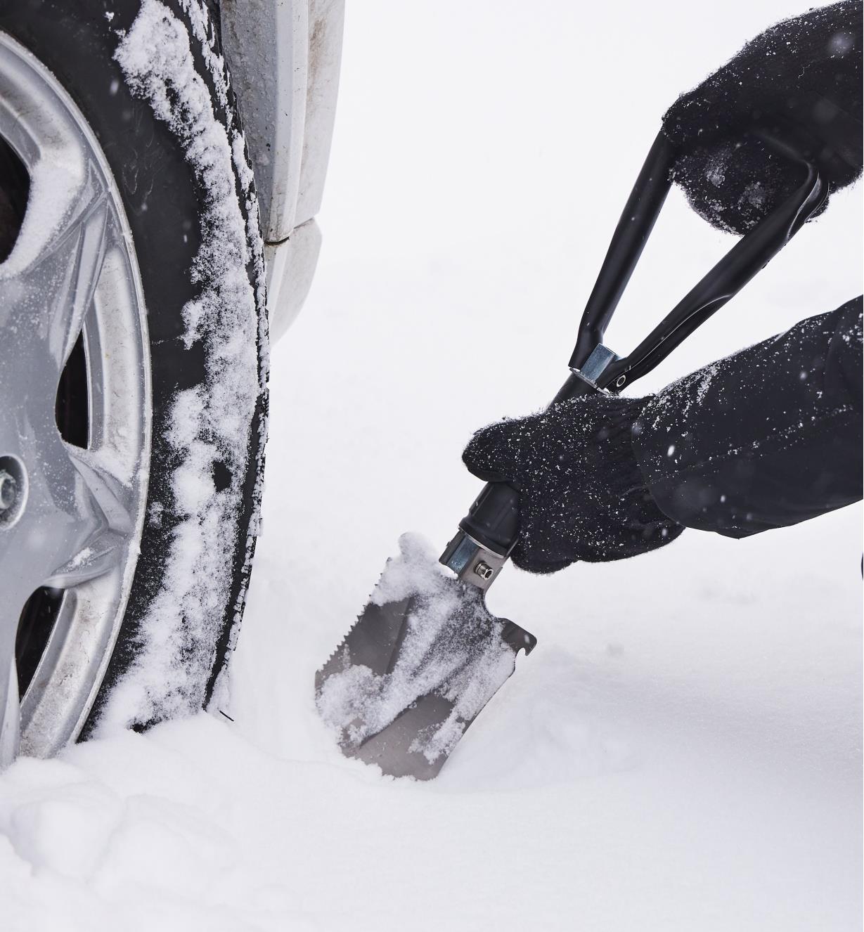 A folding shovel being used to dig snow from around a car tire