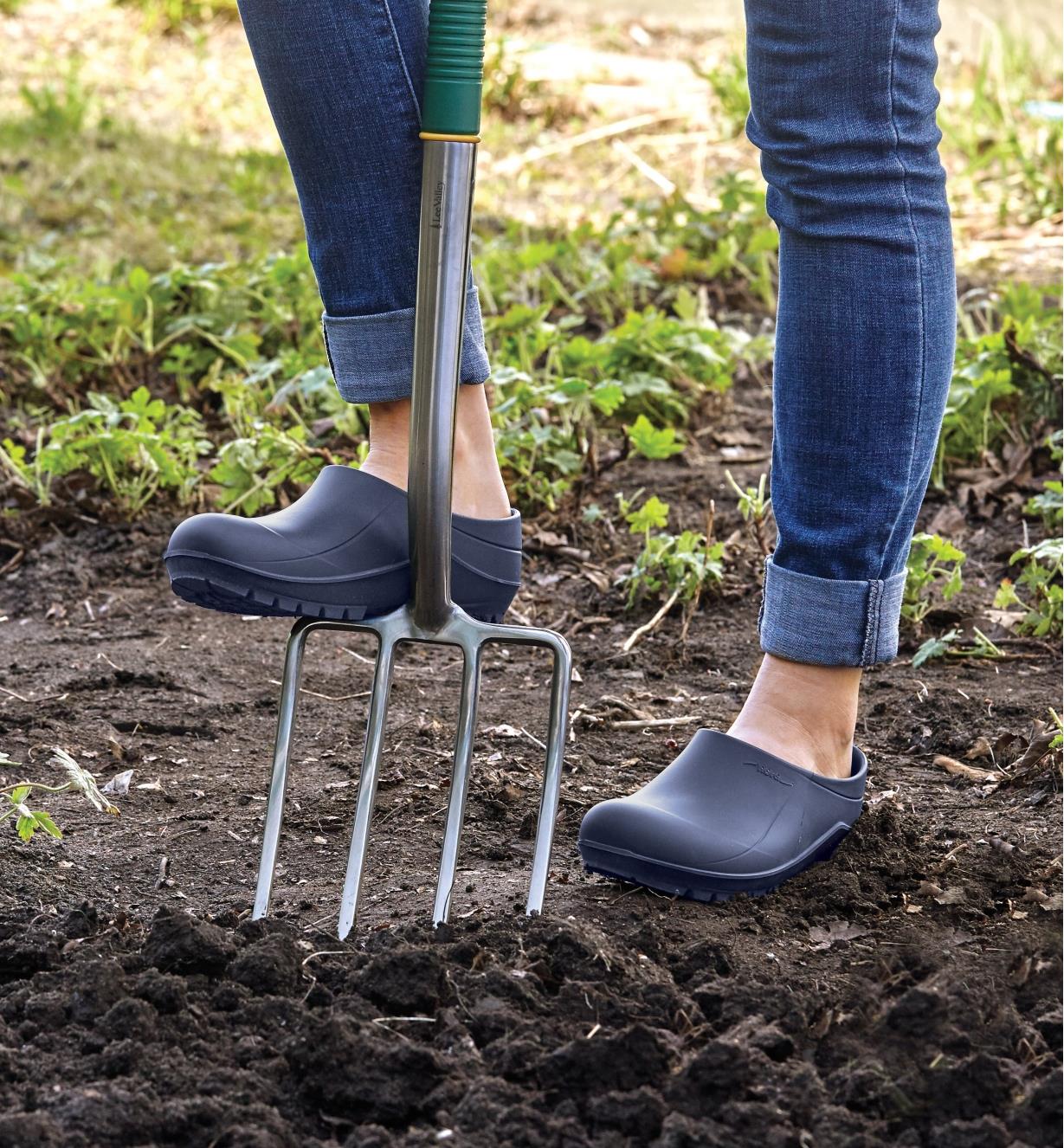 A woman wears European garden clogs while using a garden fork to loosen soil in a planting bed 
