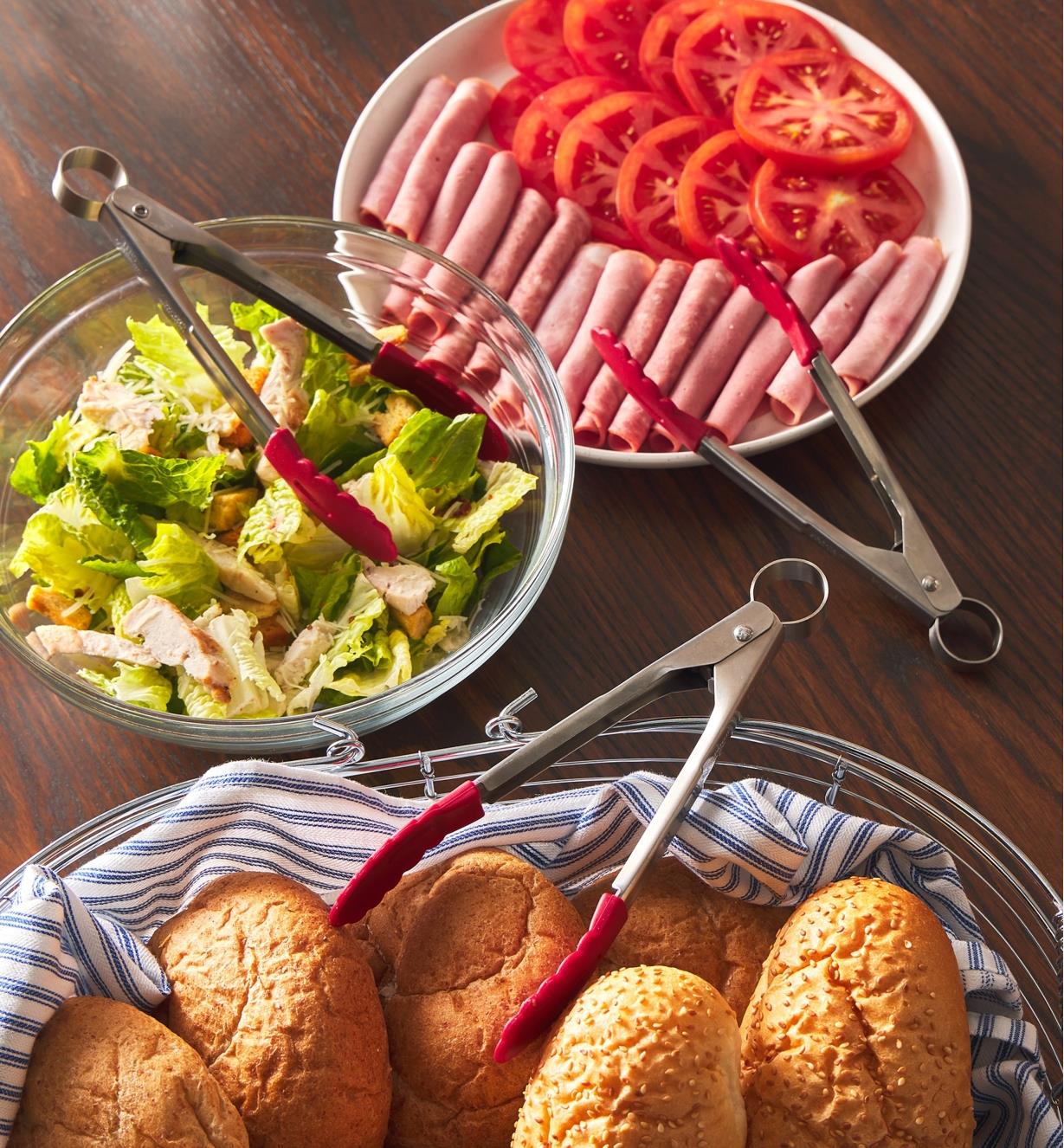 Tongs placed with rolls, a salad and a plate of meat and tomatoes