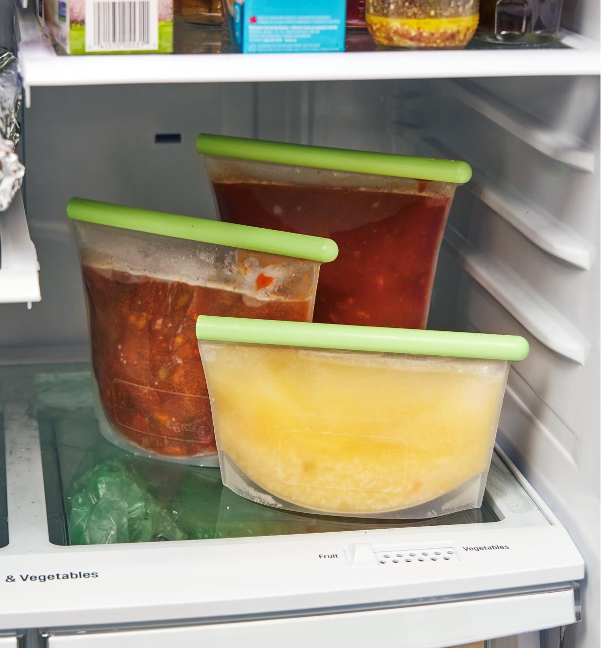 Three sizes of reusable silicone bags filled with food on a refrigerator shelf