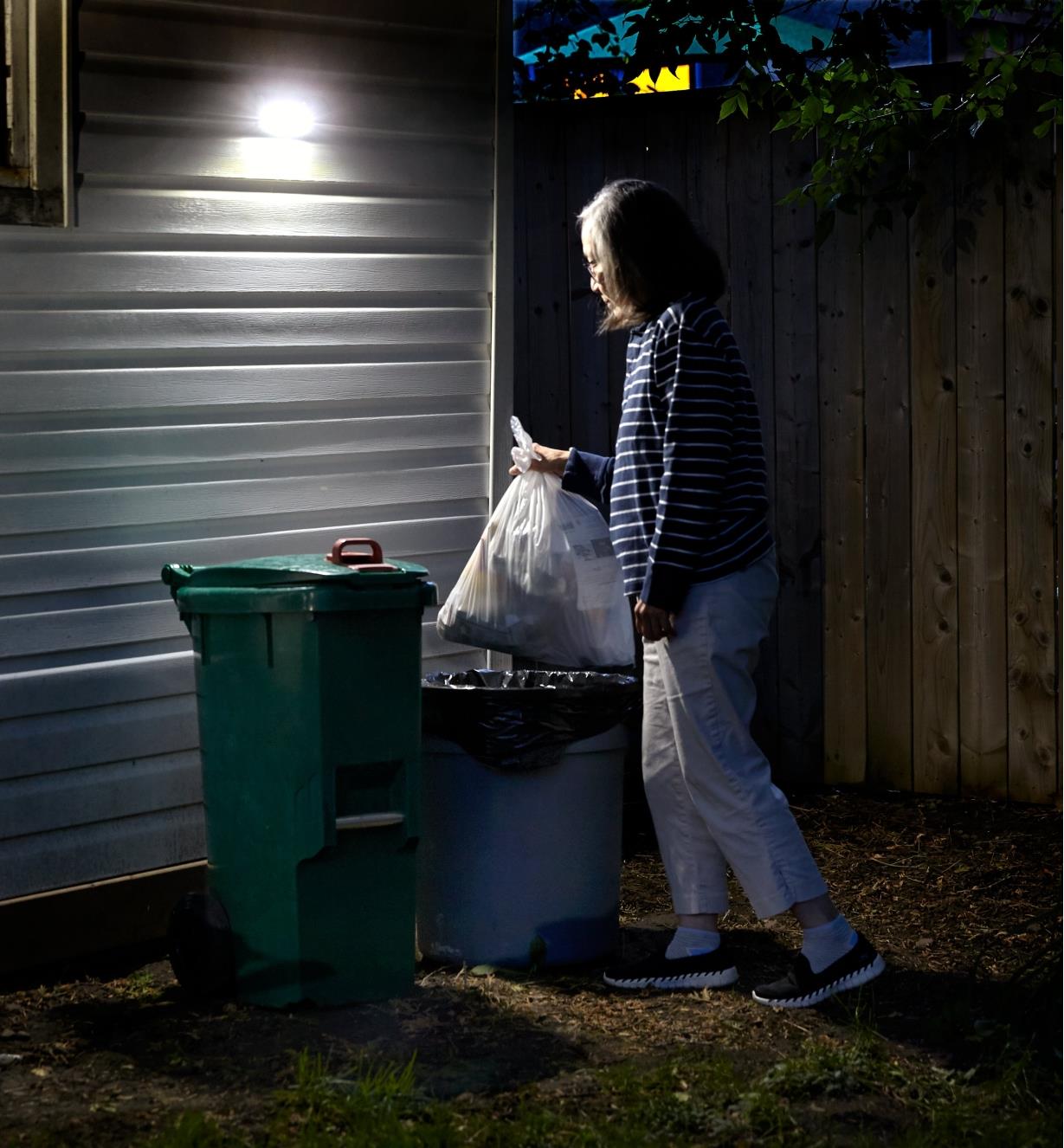 A woman discards a bag of garbage, triggering a motion-activated light mounted above the garbage can