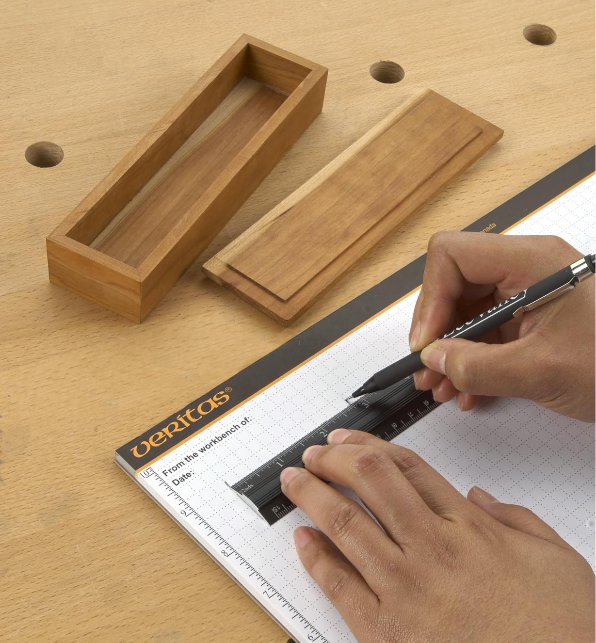 Personne utilisant une règle d'établi pour tracer une ligne sur un bloc de papier quadrillé Veritas devant une petite boîte en bois