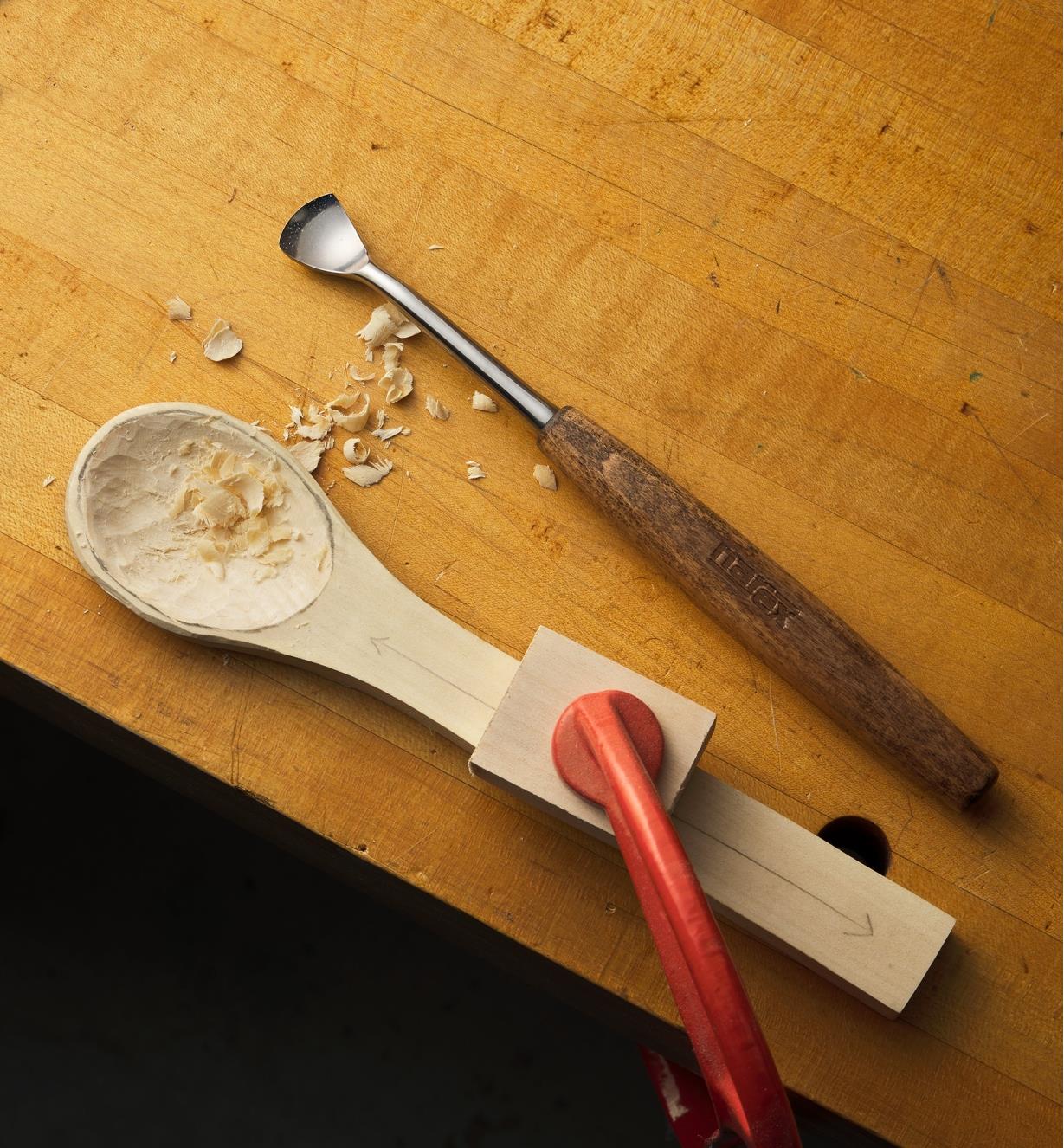 A partially finished wooden spoon clamped to a workbench and next to a carving chisel.