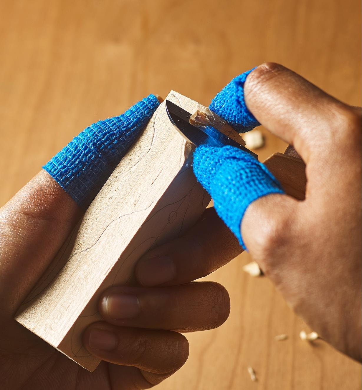 A person wearing finger guard tape beginning to carve a wood blank with a carving knife