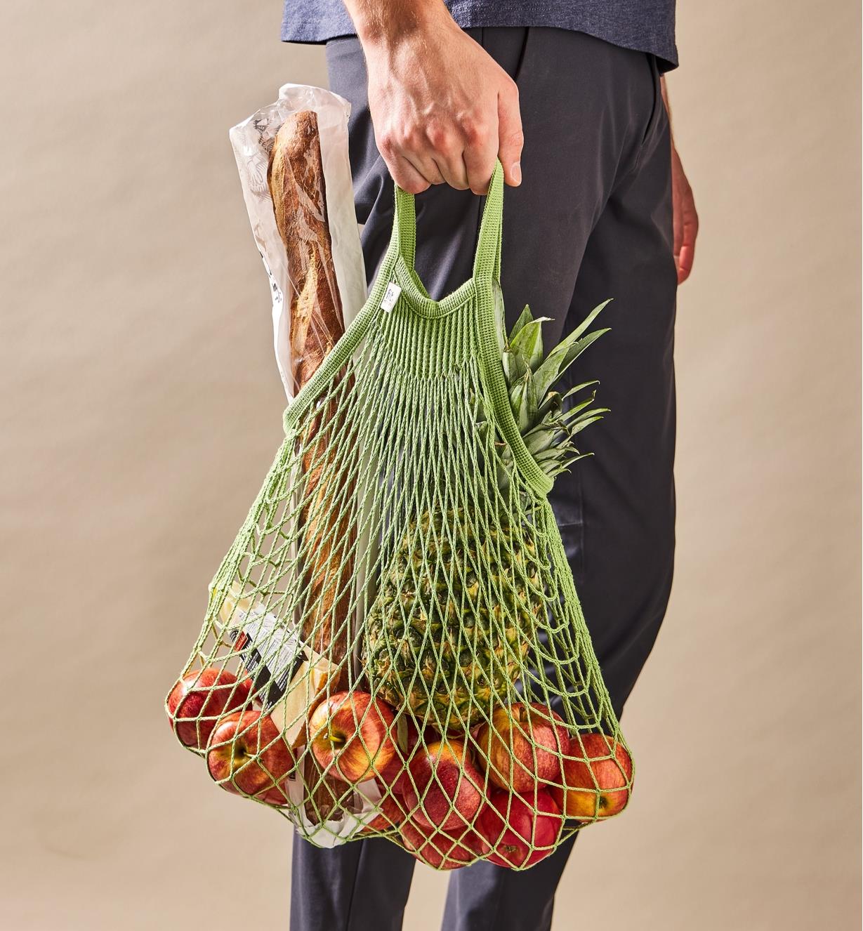 A person carrying by hand the green short-handled mesh shopping bag filled with groceries