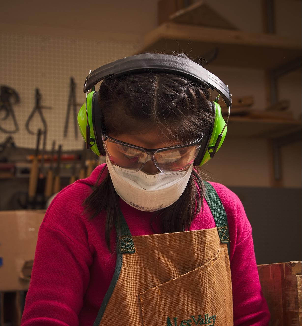 A child in a workshop wearing hearing protectors, safety glasses, a dust mask and an apron