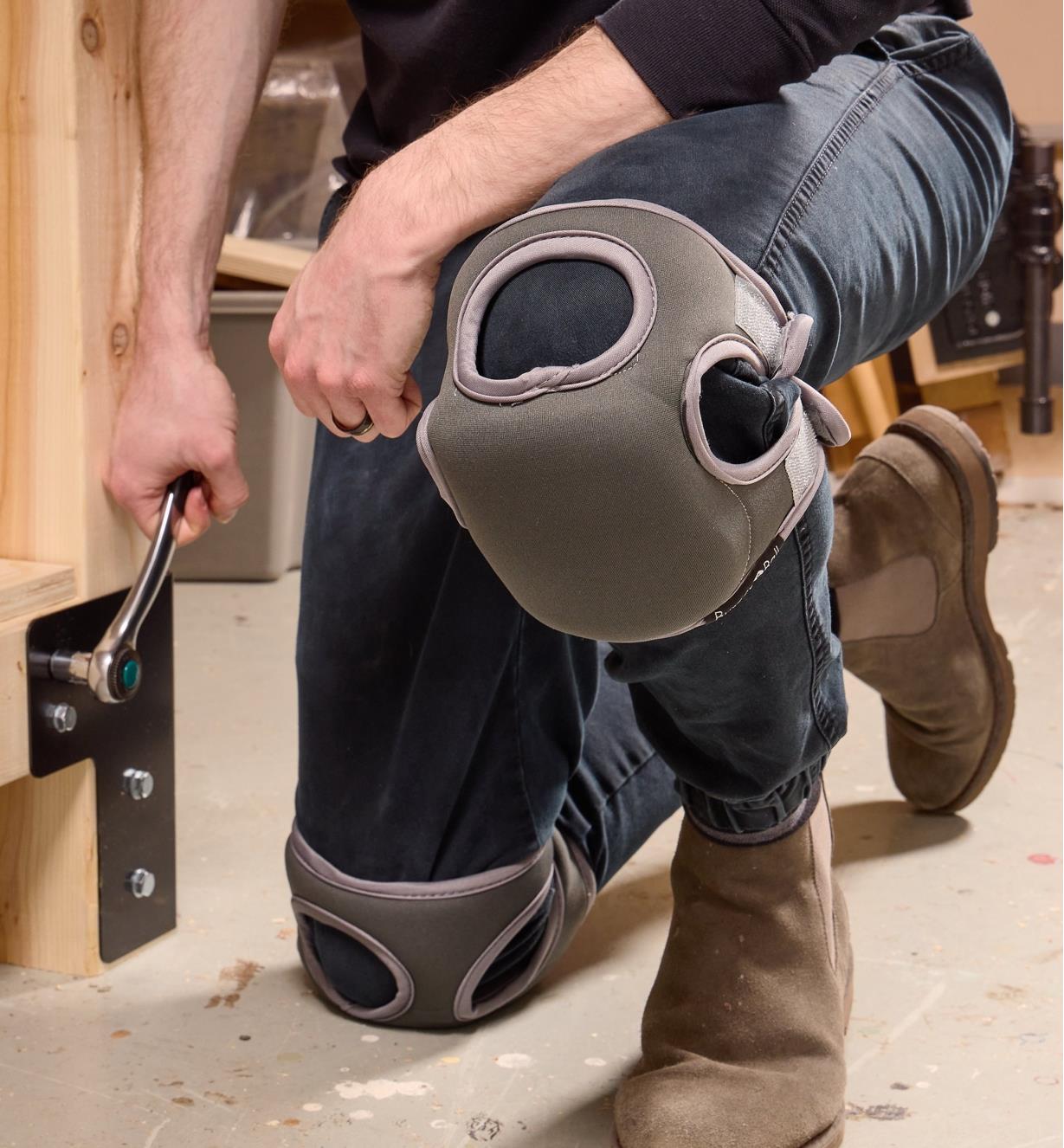 A person wearing memory foam knee pads kneels one a workshop floor while tightening workbench bolts