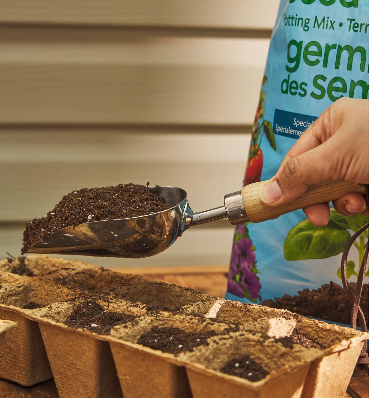 A gardener holds a stainless-steel scoop full of soil over a seedling tray