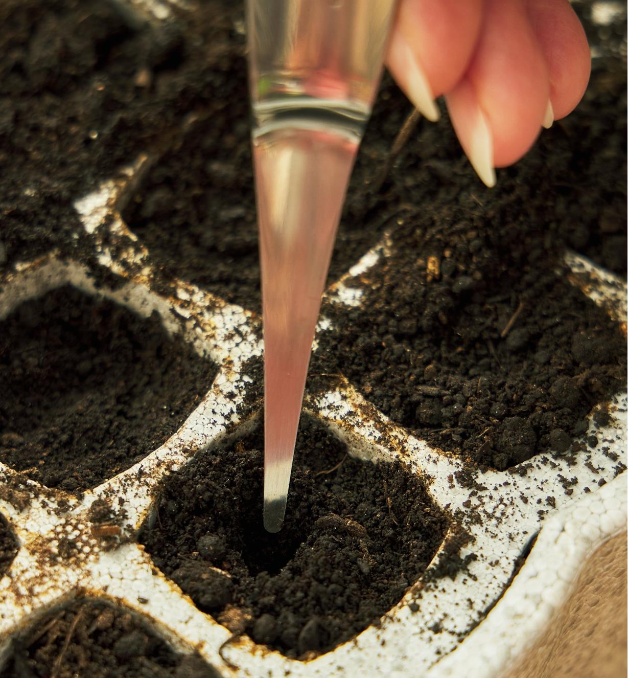 Using a potting tool to create a small hole for a seed in the soil of a seed-starter cell