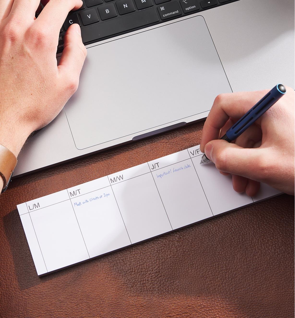 A person jots notes on a weekly tear-off planner while working on a laptop computer