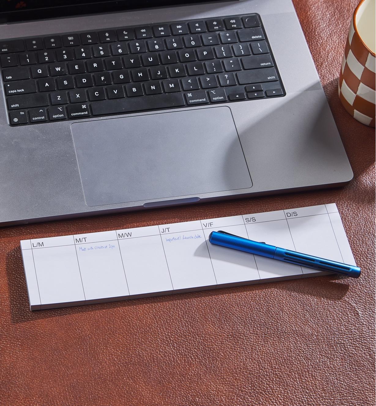 A laptop computer on a leather desk pad with a pen, mug and weekly tear-off planner with notes on it