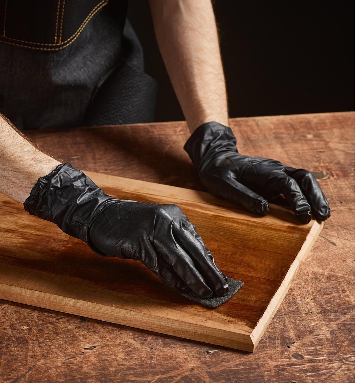 A woodworker wearing gloves uses a pad to apply Rubio Monocoat 2C to a plank of wood