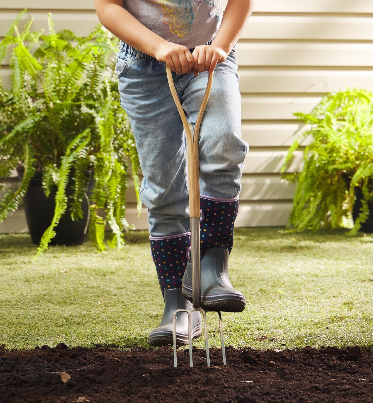 A child wearing rubber boots holds the children’s digging fork and pushes it into soil with one foot